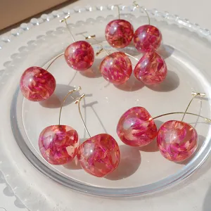 Detalle de pendientes de cereza de resina transparente con flores secas rosas y tallos dorados.