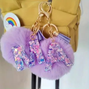 Close-up of fluffy purple pom-pom letter keychains with glitter resin and gold hardware.