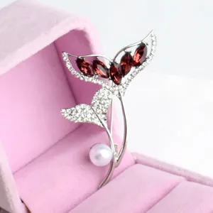 Close up view of exquisite silver fish tail brooch featuring red crystals and pearl in box.