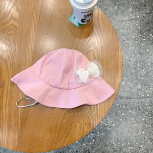 Pink infant bucket hat close-up with white lace bow and chin strap on wooden surface.
