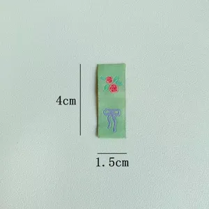 Vista dettagliata di un'etichetta in tessuto verde con rose e fiocco ricamati, misura 4x1,5cm.