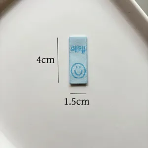 Dettaglio etichetta tessuta blu, 4x1.5 cm, motivo viso sorridente e decorazione testo Hello.