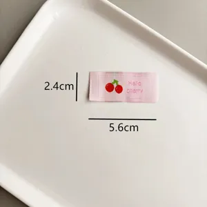 Vue détaillée de l'étiquette en tissu rose brodée de cerises avec des dimensions claires.