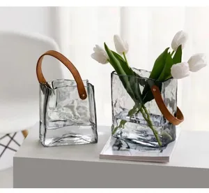 Vaso borsa in vetro trasparente con manico in pelle marrone, decorazione floreale da tavolo.