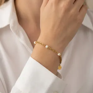 Gold-plated chain bracelet detail with small freshwater pearls and round tag on wrist.