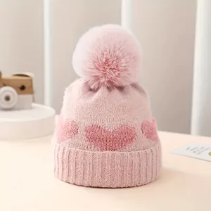 Baby knit hat detail showing pink jacquard heart pattern and soft plush pompom texture.