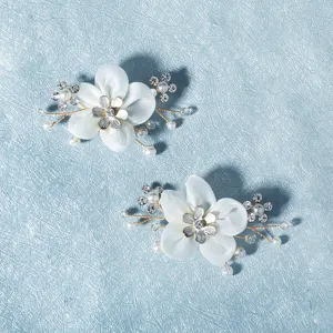 Close-up detail of white fabric flower shoe clips with pearls and rhinestones on blue backdrop.