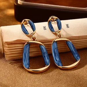 Primo piano di orecchini pendenti geometrici in smalto blu dorato con dettagli metallici intrecciati.