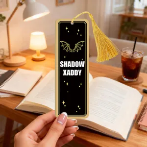Detailed view of a creative black acrylic bookmark with gold tassel, featuring bat wings and text.