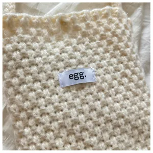 Vue détaillée d'un sac bandoulière en tricot beige douillet, avec étiquette et douce texture.