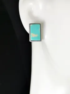 Gros plan de boucles d'oreilles clous turquoise rectangulaires sur présentoir blanc.
