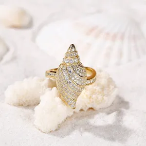 Primo piano dettaglio di un elegante anello conchiglia dorato con strass su sfondo sabbia fine.