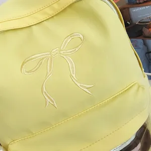 Zaino giallo con delicato ricamo a fiocco, ideale per l'uso scolastico quotidiano degli studenti.