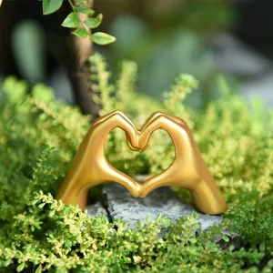 Close-up detail of a modern gold heart hand gesture sculpture ornament placed among lush green plants.
