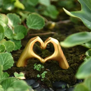 Close-up of golden heart gesture statue ornament in natural moss and green leaves scene.