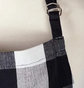Close up view of black and white plaid apron fabric, adjustable shoulder strap with metal ring.