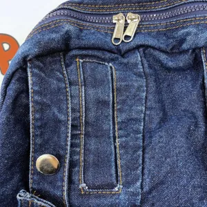 Denim backpack detail view, dark blue fabric with contrast stitching and metal hardware.
