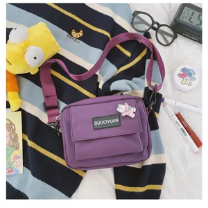 Vista detallada del bolso cruzado de lona morada con bolsillo delantero y etiqueta elegante.