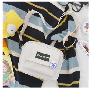 Vista detallada de un bolso cruzado blanco minimalista con bolsillo delantero y etiqueta.