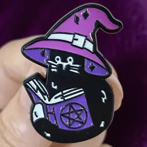Close-up detail of a cute gothic black cat enamel pin wearing a purple witch hat reading book.