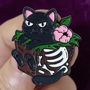 Close-up detail shot of a cute dark style black cat enamel pin with skeleton bones and flower.