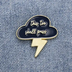 Enamel cloud lightning pin detail, black and gold metal badge showing this too shall pass.