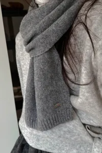 Nahaufnahme des grauen Damen Strickschals im Winter, verziert mit einem kleinen runden Metallschild.