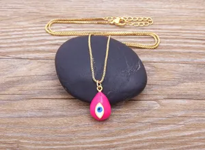 Close-up detail of a gold-plated necklace with a pink drop evil eye pendant on a smooth stone.