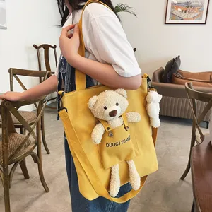 Vista detallada del bolso cruzado de lona amarilla con bolsillo de oso de peluche y colgante.