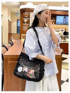 Detail shot of black ita bag with clear window showing anime badges, lifestyle cafe setting.