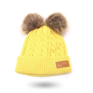 Close-up of yellow kids knit beanie detail, featuring fluffy faux fur pom poms and leather tag.