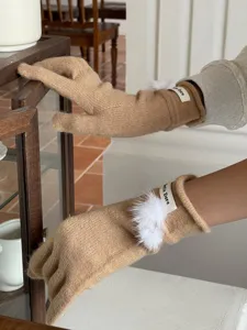 Nahaufnahme von kuscheligen Kamel-Strickhandschuhen mit süßem weißem Plüschball und Markendetail.