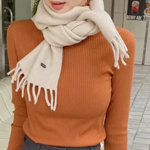 Dettaglio in primo piano di morbida sciarpa in maglia beige con frange ed etichetta metallica.