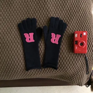 Nahaufnahme schwarzer Strickhandschuhe mit rosa R-Buchstabe auf braunem Stoffgrund.