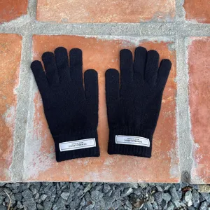 Detailaufnahme schwarzer Strickfünffingerhandschuhe mit weißem Markenetikett am Bündchen.