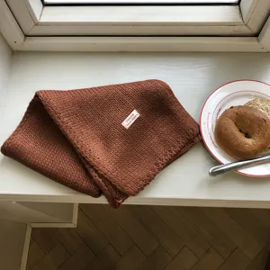 Gemütlicher brauner Strickschal in warmer Detailansicht auf einer weißen Fensterszene.
