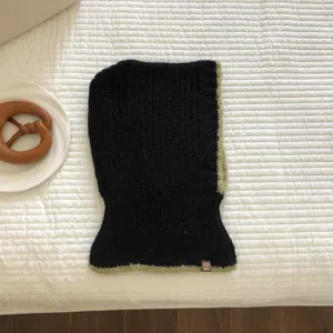 Detail view of a black knitted balaclava winter hat with green contrast edge and woven tag.