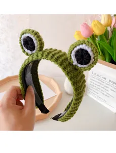 Close-up detail shot of the green knitted frog headband featuring cute big eyes, soft wool texture.