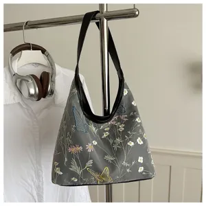 Vista detallada del bolso de hombro gris bordado con flores y mariposas con correa de cuero negro.