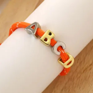 Accesorio de pulsera con alfabeto DIY, cuentas de letras doradas y plateadas en cuerda naranja tejida.