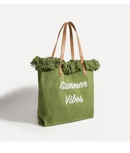 Vista detallada del bolso tote de lona verde con bordado blanco y flecos decorativos.