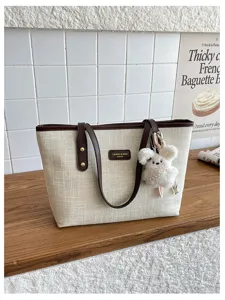 Minimalist beige canvas tote bag detail view, featuring brown leather handles and cute plush pendant.