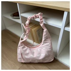 Pink gingham shoulder bag with white bow embroidery, detailed view on white shelf background.