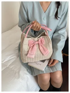 Close-up detail of a straw tote bag with a pink bow held by a model in a bedroom setting.