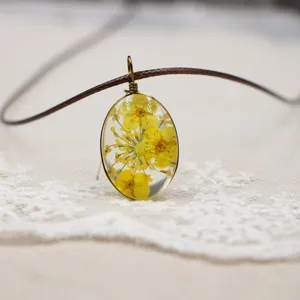 Primer plano de un colgante ovalado de resina y flores secas con collar de cordón marrón.