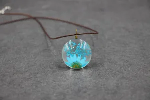 Close-up detail of dried flower glass pendant necklace with blue daisy on dark grey background.