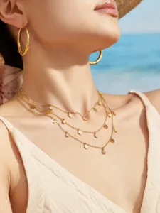 Detailaufnahme mehrlagiger Goldketten mit kleinen Anhängern und Ohrringen am Strand.
