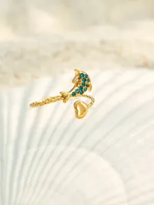 Detailansicht eines goldenen Delphin-Offenrings mit blauen Zirkoniasteinen auf einer weißen Muschel