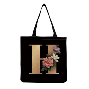 Black canvas tote bag featuring a gold floral monogram letter H design for daily shopping use.