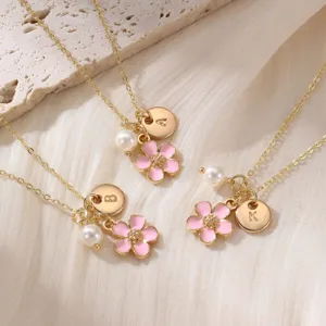 Détail du collier doré avec pendentif fleur en émail rose, perle et breloques lettres.